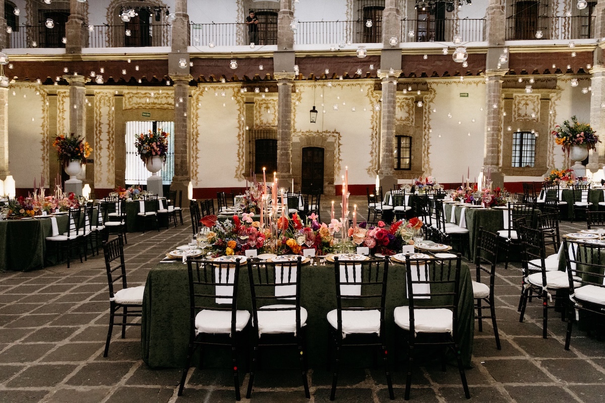 Elegant banquet hall with stone walls, round and rectangular tables set for an event, floral centerpieces, candles, and string lights overhead.
