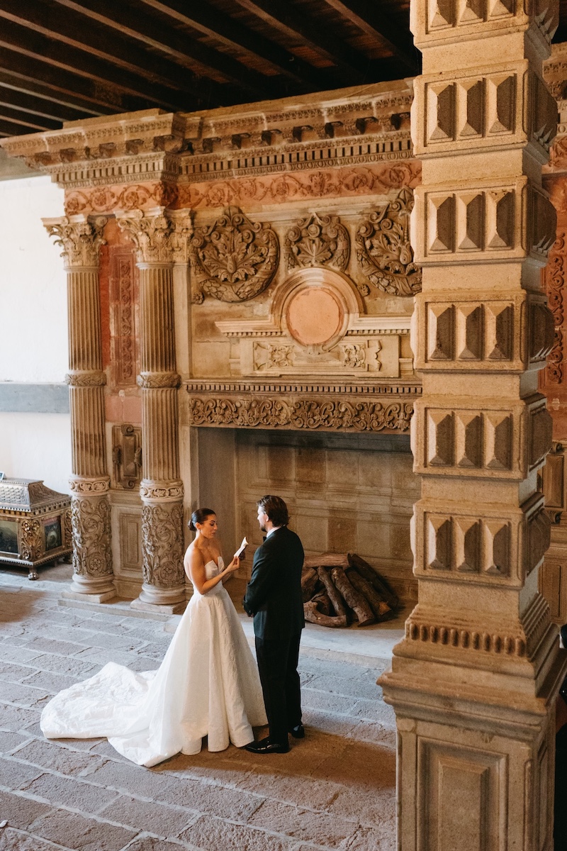 A bride and groom stand facing each other in formal attire inside an ornate room with carved stone pillars and an elaborate fireplace.