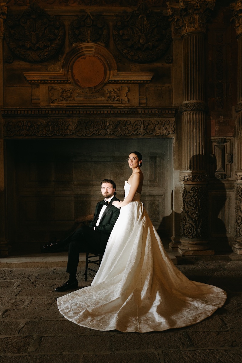 A bride in a white gown stands beside a seated groom in a tuxedo, posed in front of an ornate, dimly lit, historical backdrop.