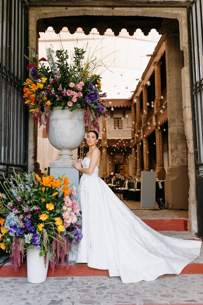 A bride in a white wedding dress stands beside large floral arrangements in an outdoor courtyard decorated with string lights.