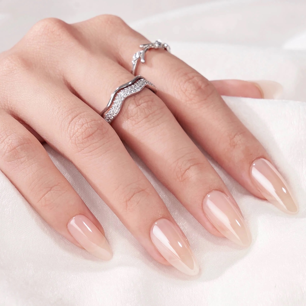 A hand with long, almond-shaped nails painted in a glossy nude shade, resting on white fabric and adorned with two silver rings.