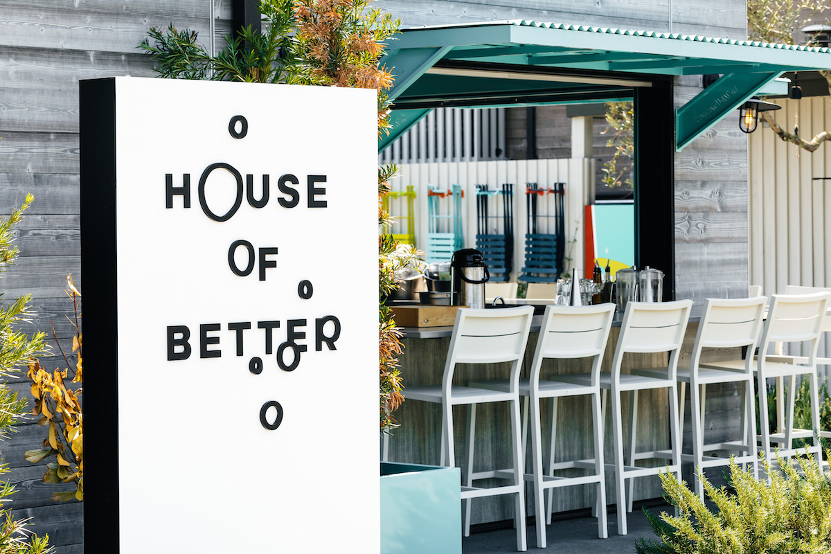 Outdoor bar area with white stools lined up at a counter; a modern sign in the foreground reads "House of Better.