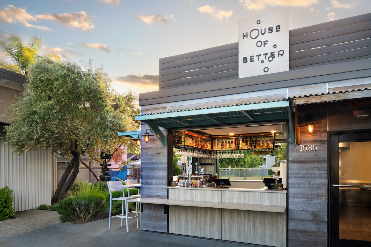 Small outdoor café with a service window, labeled "House of Better," surrounded by greenery under a partly cloudy sky.