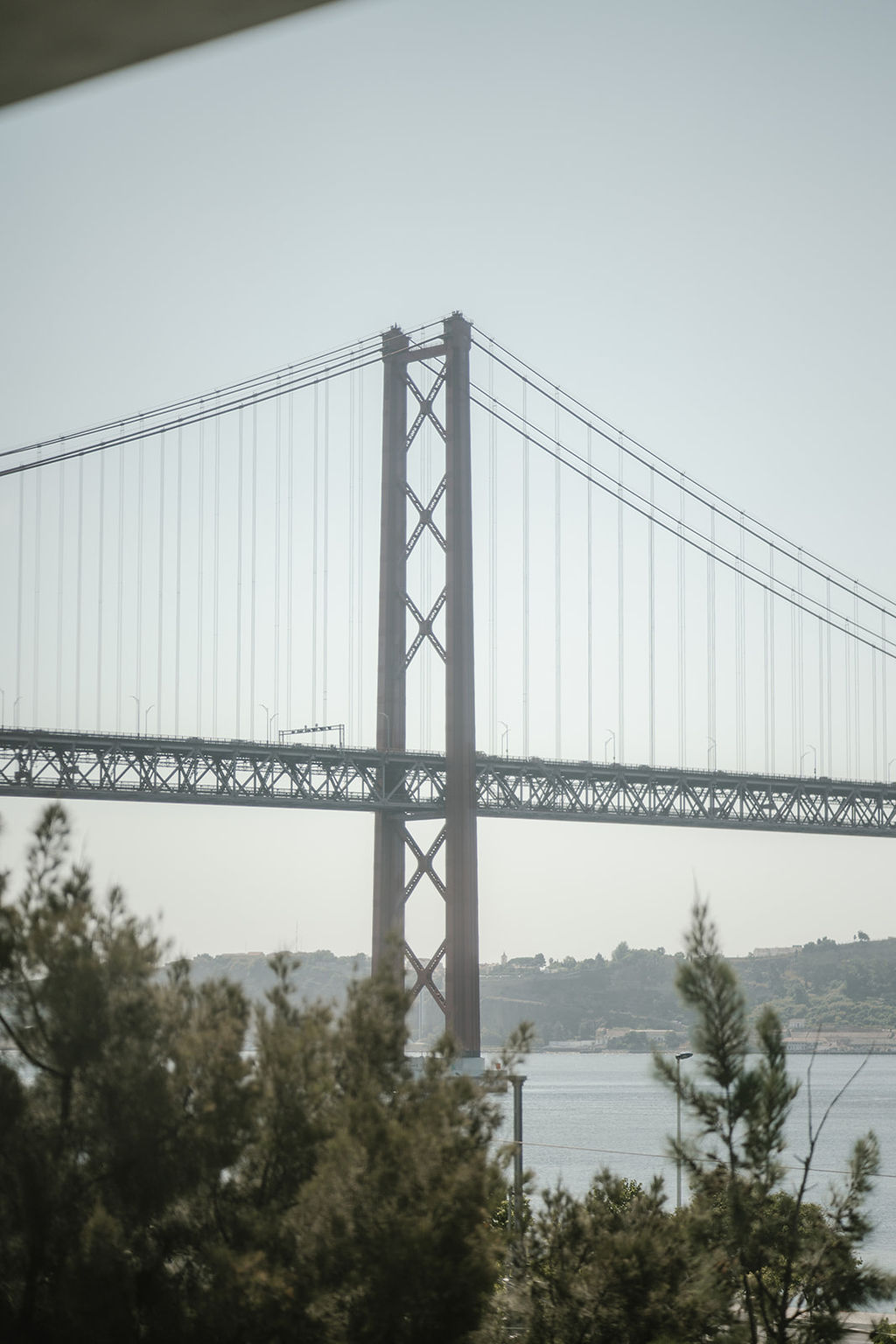 A suspension bridge with red towers spans across a body of water, with trees and foliage in the foreground.