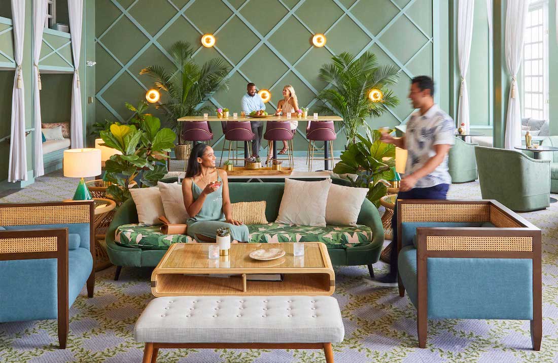 Modern lounge with people sitting and standing, tropical decor, green walls, and a bar in the background.
