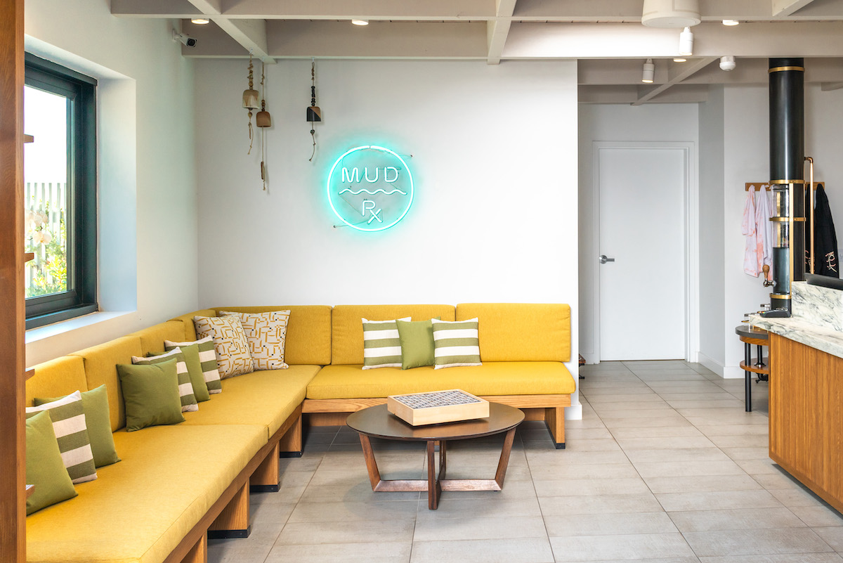 Modern lounge area with yellow sectional sofa, green and white pillows, a round coffee table, and a neon “MOC” sign on the wall.