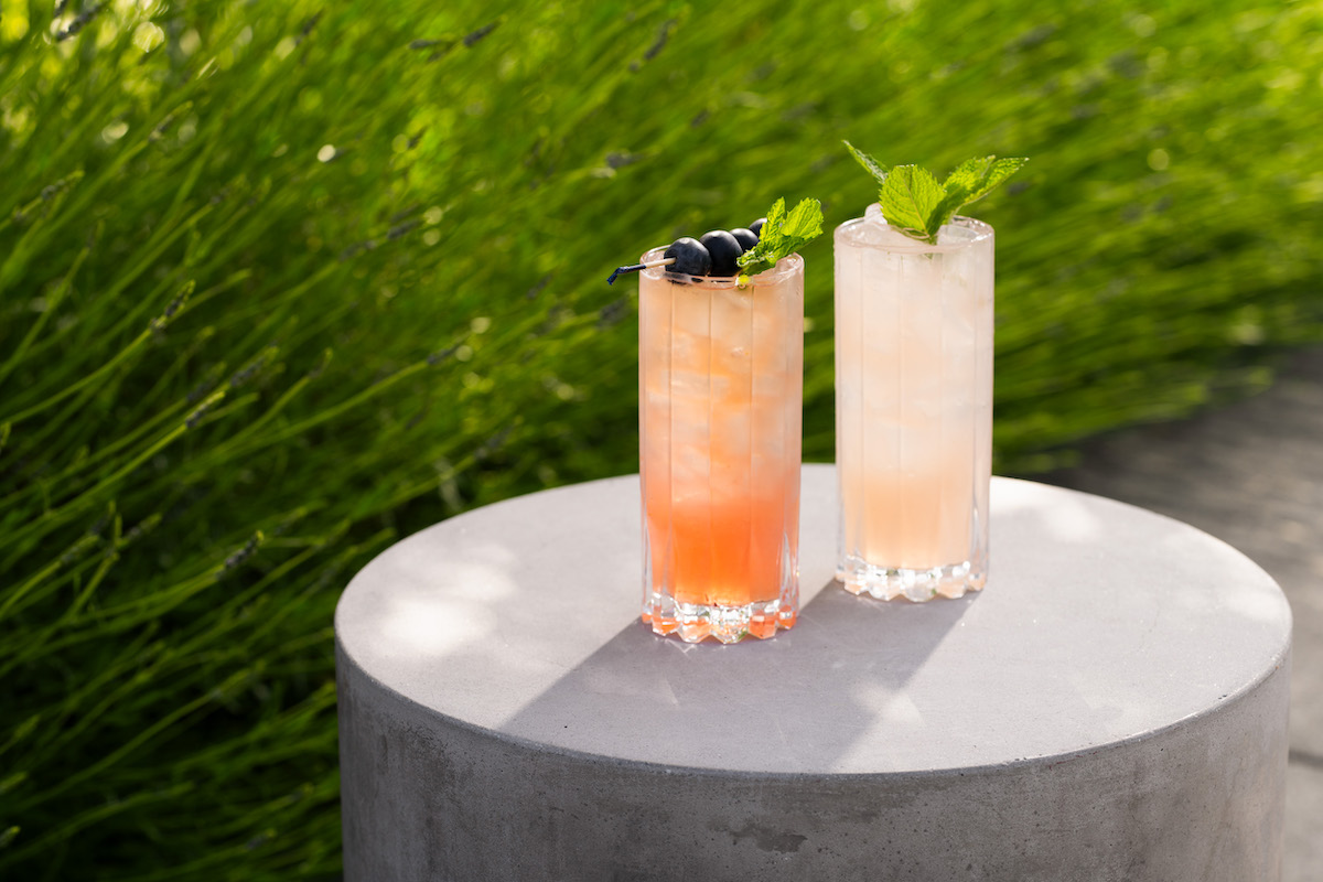 Two tall glasses of iced drinks, one pink-orange with berries and one pale with mint, sit on a round concrete table outdoors with green foliage in the background.