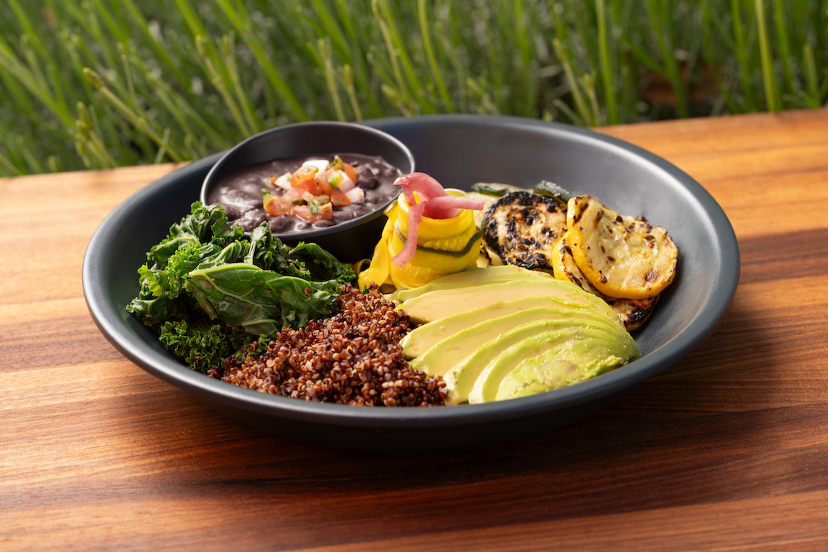 A black bowl with sliced avocado, red quinoa, kale, grilled vegetables, and a small serving of black beans with salsa, set on a wooden table.