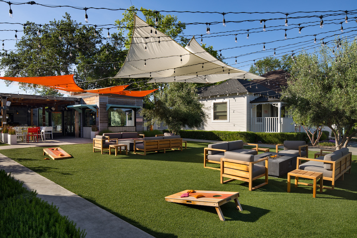 Outdoor patio area with modern wooden furniture, string lights, shade sails, and cornhole boards set up on artificial grass near a house.