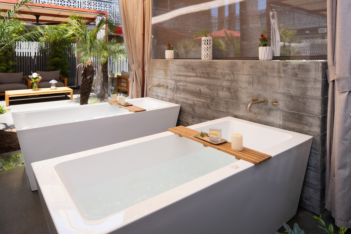 Two white bathtubs filled with water are placed side by side outdoors, with wooden trays holding candles and towels, next to a concrete wall with a faucet.