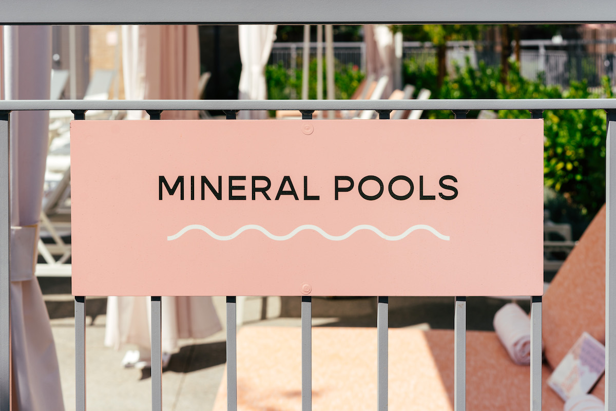 A pink sign with the words "MINERAL POOLS" and a wavy line is attached to a metal fence; lounge chairs and towels are visible in the background.