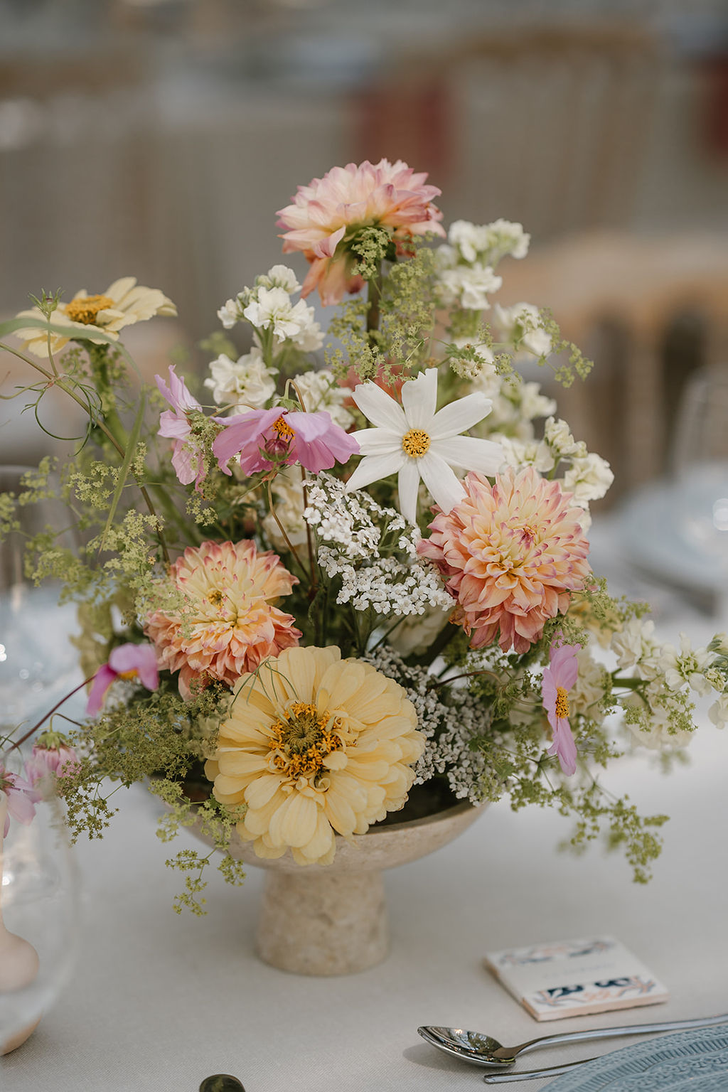 A floral arrangement in a small vase featuring yellow, pink, and white flowers with green foliage, placed on a white tablecloth.
