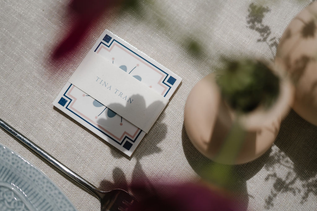 A square tile as a place card with blue accents lies on a linen tablecloth beside a glass plate, spoon, and a blurred vase with flowers.