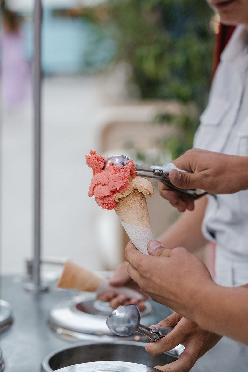 A person is scooping pink ice cream onto a cone held by another person at an outdoor ice cream stand.