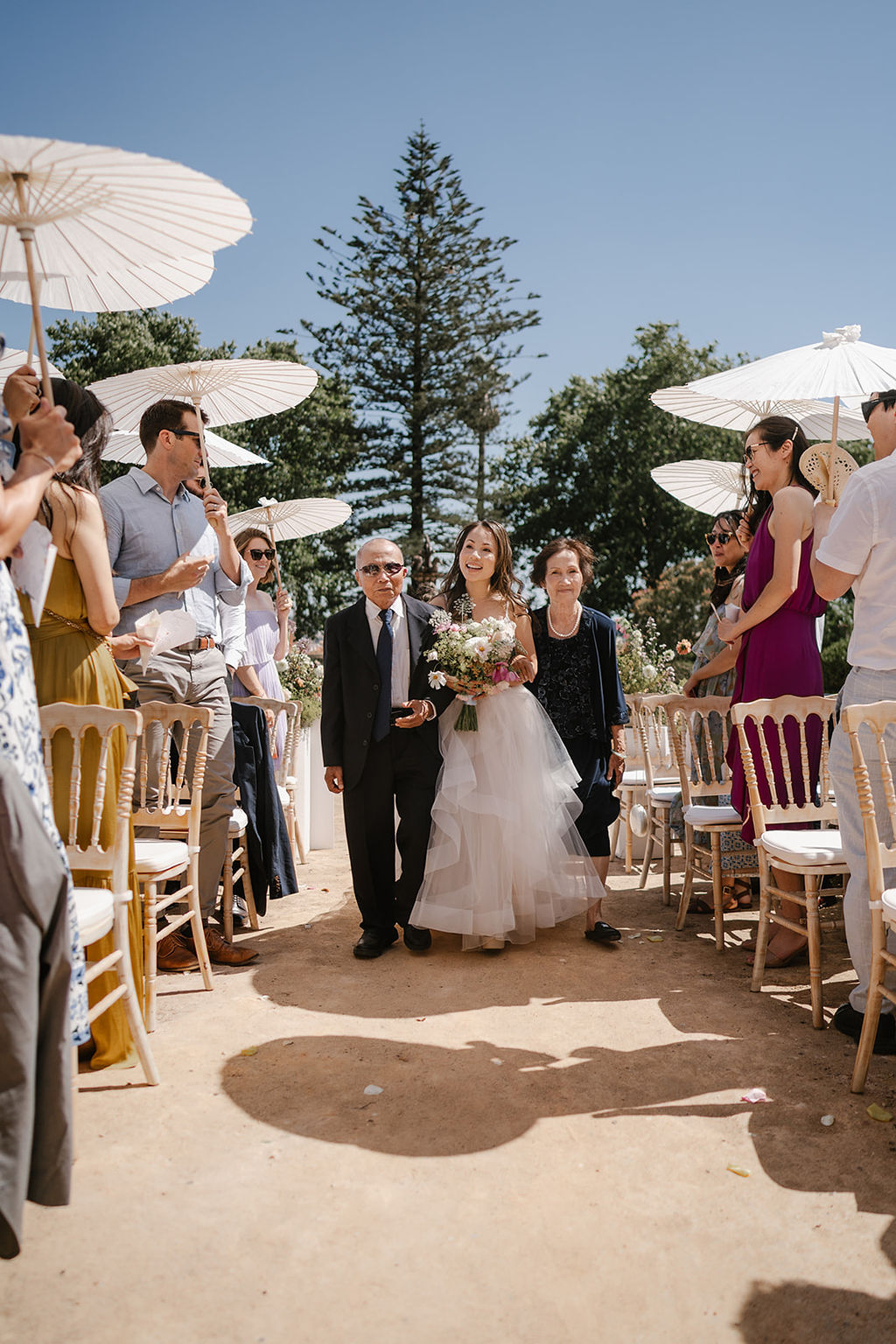 A bride walks down an outdoor aisle holding a bouquet, accompanied by two people, as guests stand and watch under white umbrellas.