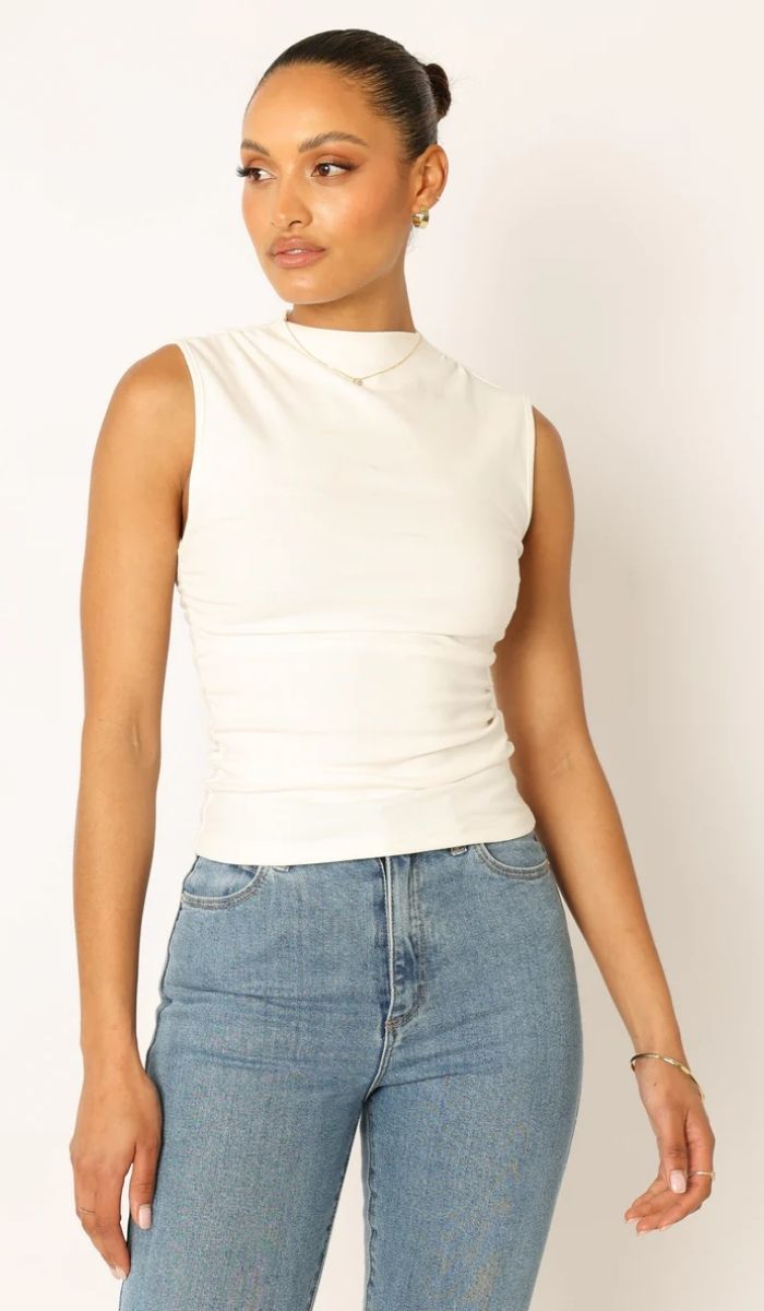 Woman wearing a sleeveless white top and blue jeans stands against a plain light background, looking slightly to the side.