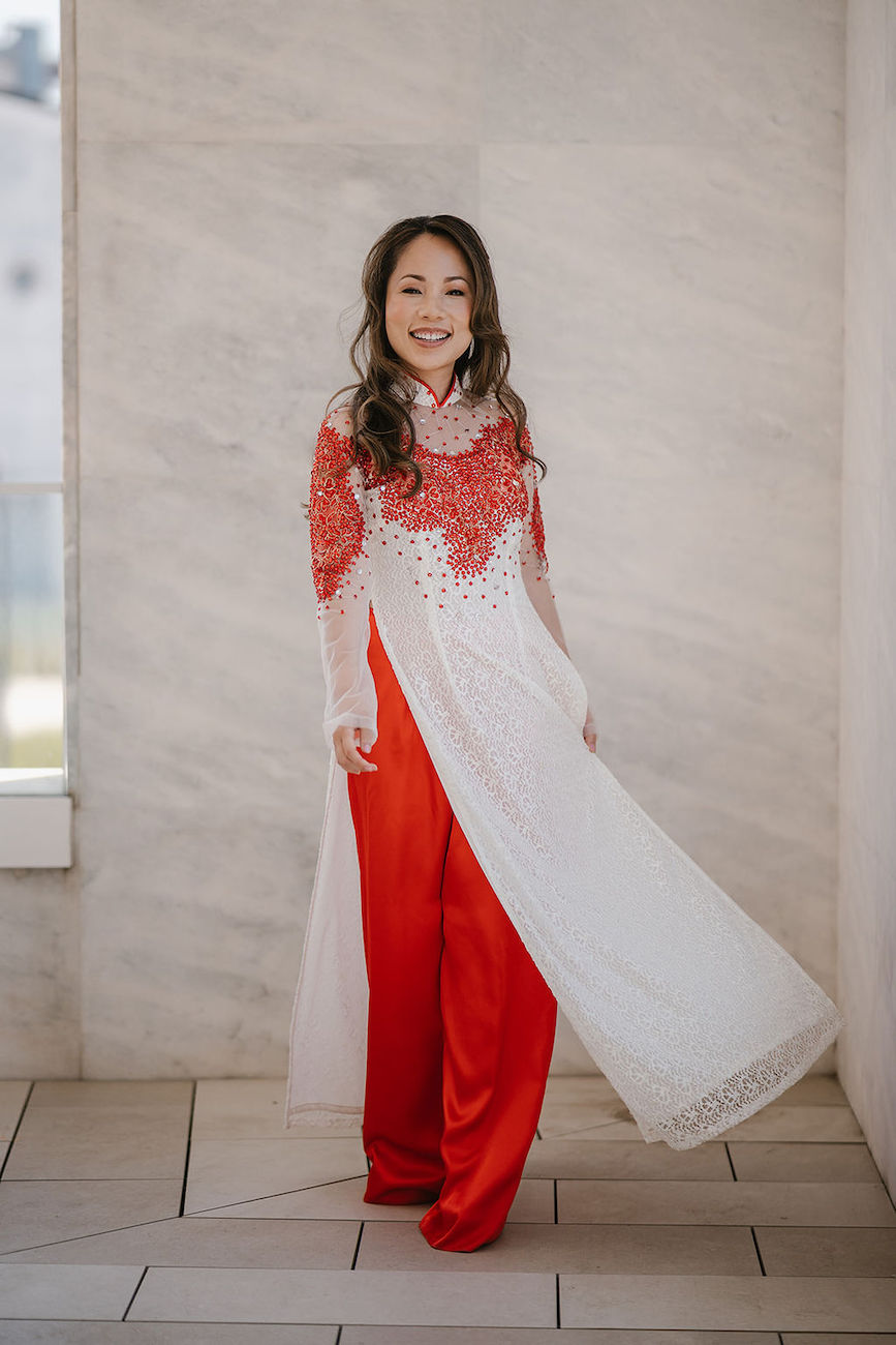 A woman stands indoors wearing a traditional outfit with a white and red long tunic over red pants, smiling at the camera.