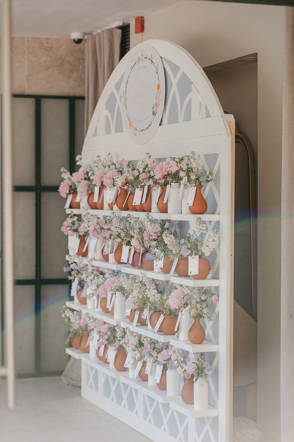 A white arched shelf displays rows of small vases filled with pink and white flowers, each vase holding a card.