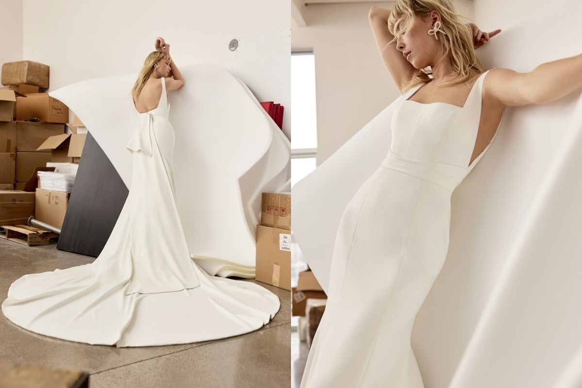 A woman in a fitted white gown poses indoors against large white panels, surrounded by cardboard boxes.