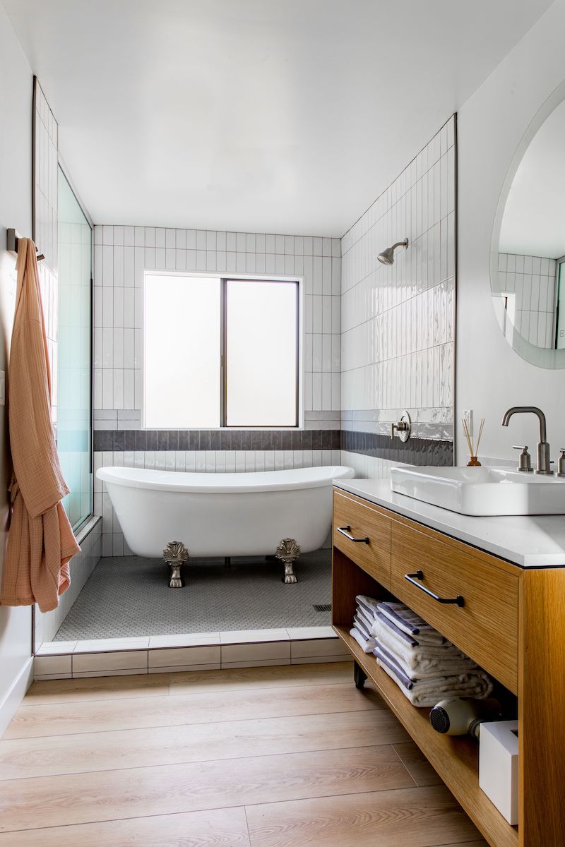 Modern bathroom with a clawfoot bathtub, wood vanity with sink, round mirror, and shelves holding towels and books; tiled walls and wood flooring.