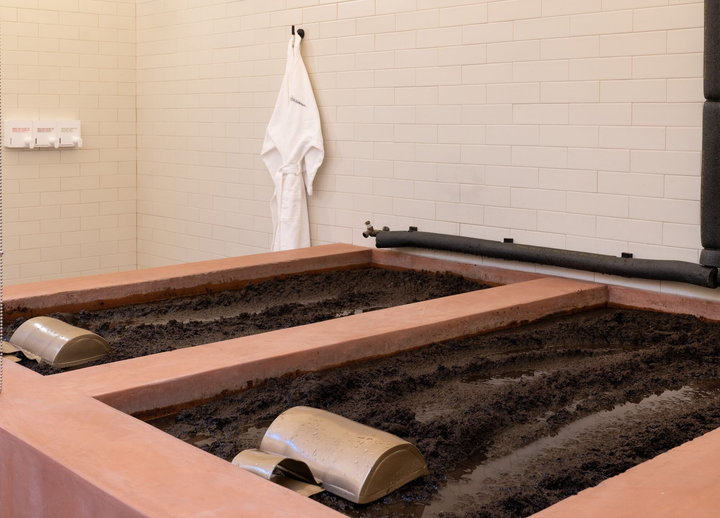 A spa room with two rectangular mud baths, each with a metal headrest, tiled walls, and a white robe hanging on a hook.