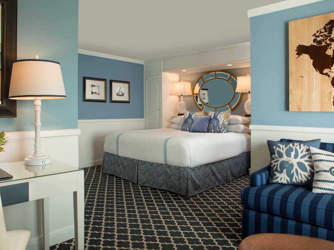 A neatly made bed with blue and white bedding sits in a hotel room with blue walls, patterned carpet, a desk, and a blue striped armchair with decorative pillows.