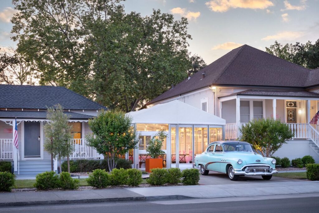 A vintage blue car is parked in front of a white house with a tented outdoor seating area; trees and shrubs line the sidewalk under a partly cloudy sky.