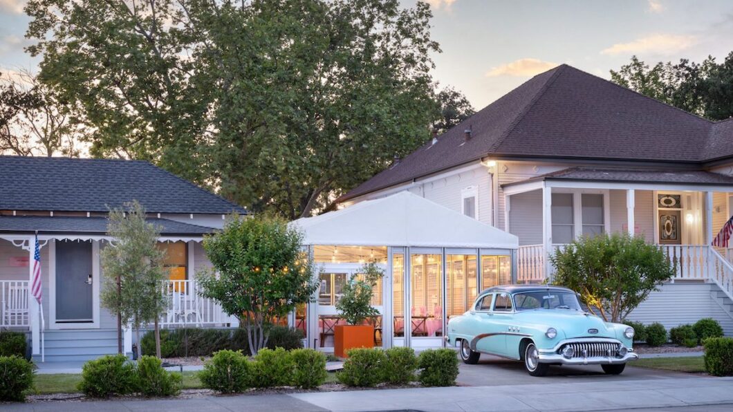 A vintage blue car is parked in front of a white house with a tented outdoor seating area; trees and shrubs line the sidewalk under a partly cloudy sky.