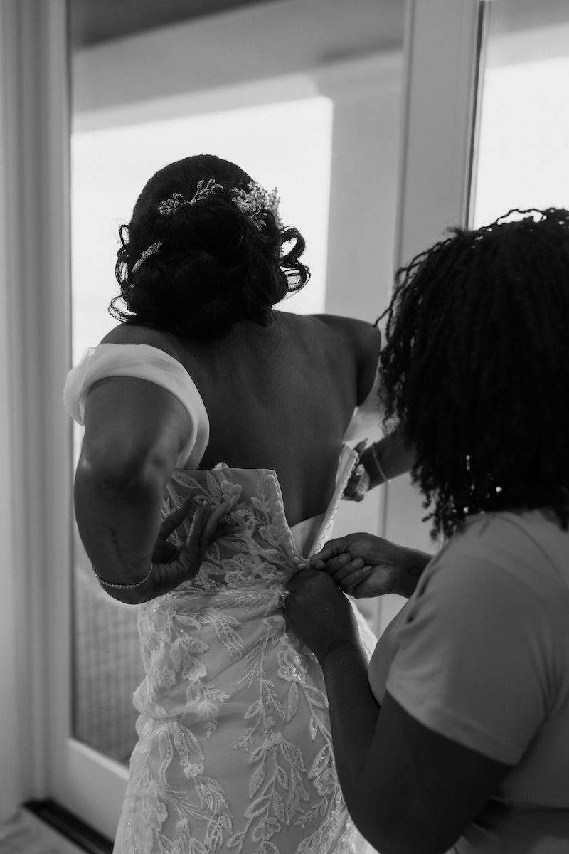 A woman helps another woman zip up the back of her wedding dress in front of a glass door.