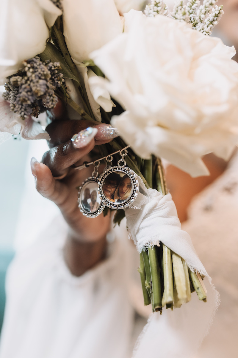 A hand holds a bouquet of white flowers with two photo lockets attached to the stems, wrapped in white fabric.