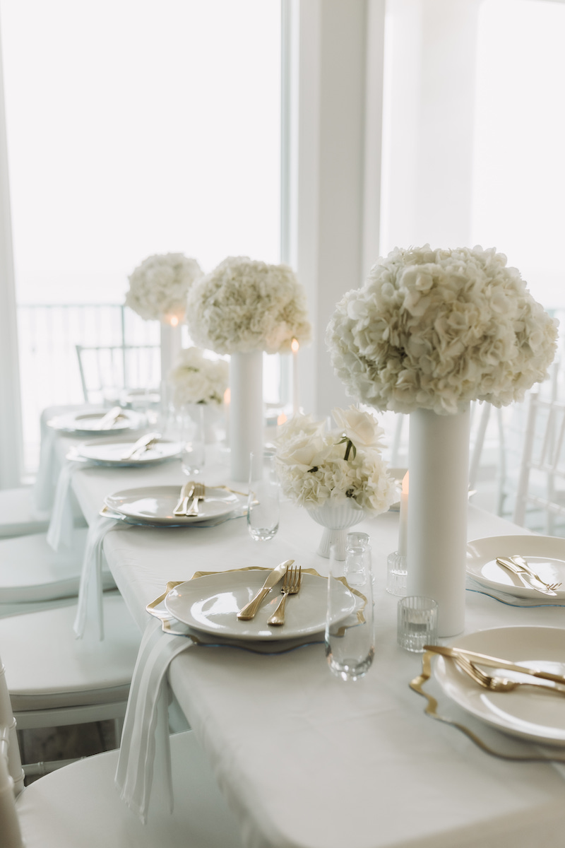 A white table is elegantly set with white plates, gold cutlery, and tall vases holding white flower arrangements in a bright, airy room.