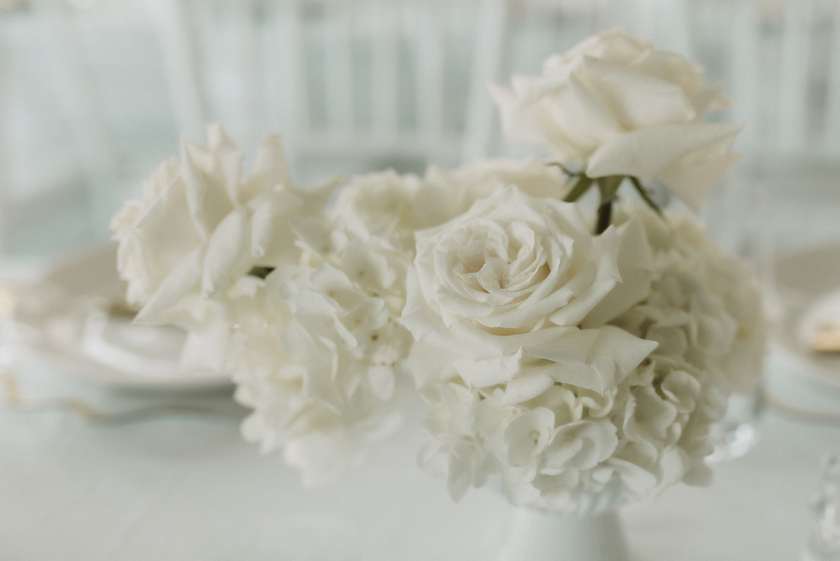 A close-up of white roses and carnations arranged in a white vase, with a blurred background of white chairs and table settings.