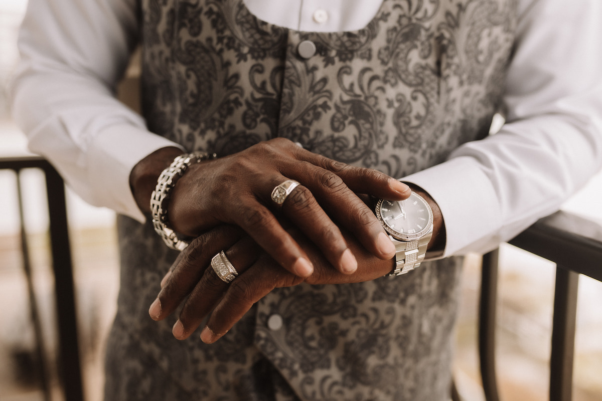 A person wearing a patterned gray vest, white shirt, multiple rings, a bracelet, and a watch, with their hands folded on a railing.