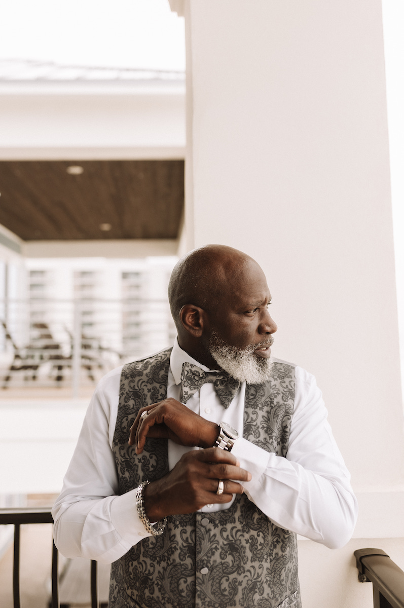 A man with a gray beard, wearing a patterned vest, white shirt, and bow tie, stands indoors adjusting his sleeve and looking to the side.