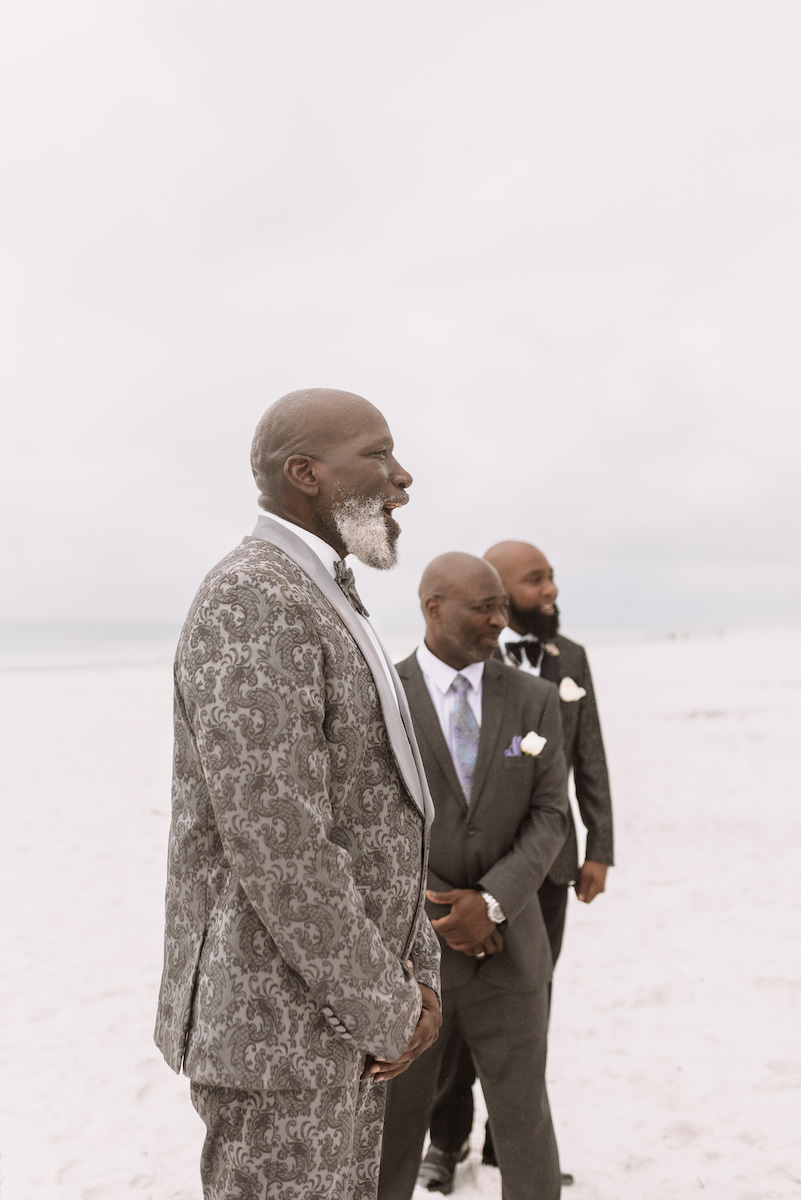 Three men in suits stand on a sandy beach, facing left. The sky is overcast and the background shows an empty shoreline.