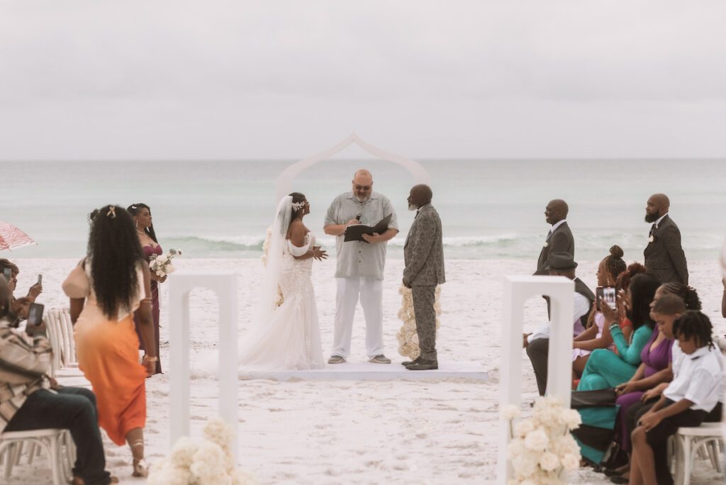 A bride and groom stand with an officiant under an arch on a sandy beach, surrounded by seated and standing guests, with the ocean in the background.