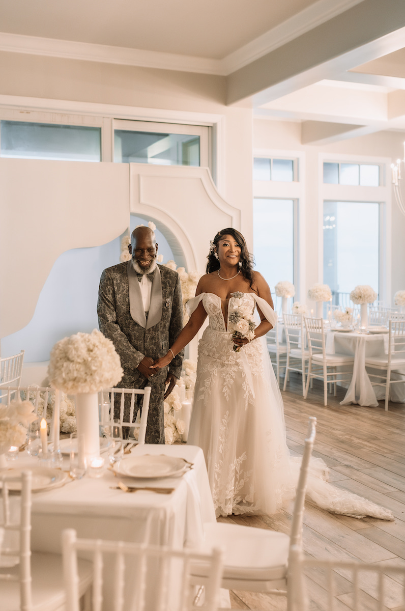 A bride in a white gown stands holding hands with an older man in a suit inside a decorated wedding reception venue with white flowers and chairs.