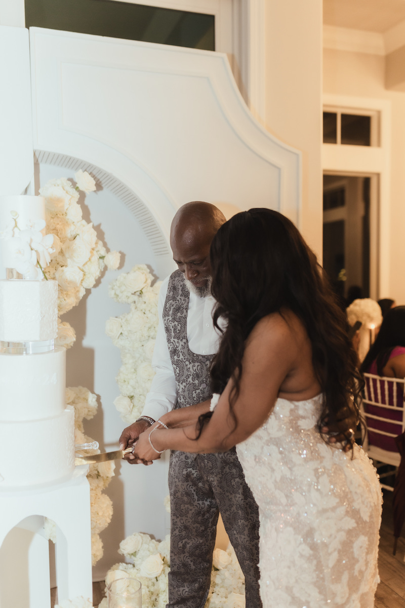 A couple in formal attire cuts a white tiered cake together at an indoor event decorated with white flowers.