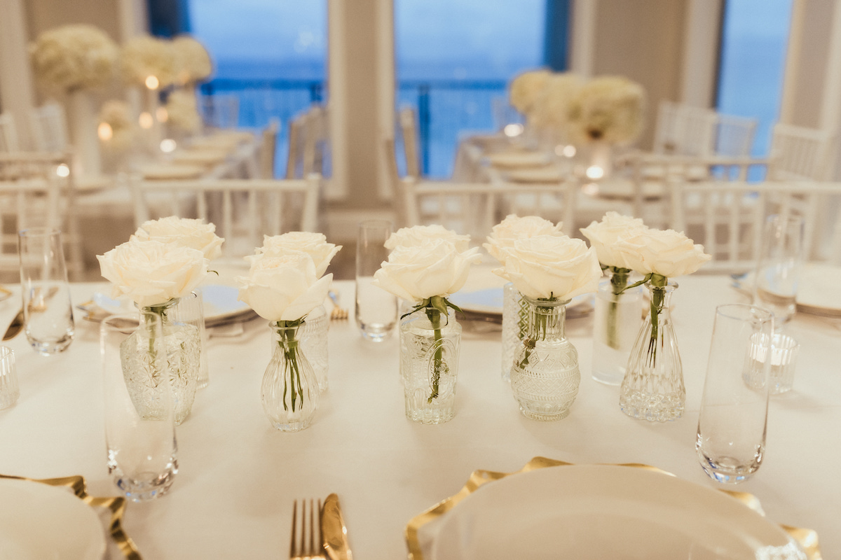 A close-up of a formal dining table set with white plates, gold-edged chargers, and small glass vases holding white roses. White chairs and more floral arrangements are in the background.