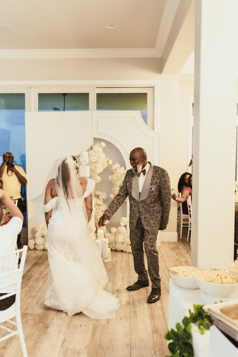 A bride and groom share their first dance in a bright, decorated indoor wedding venue, while guests watch and take photos.
