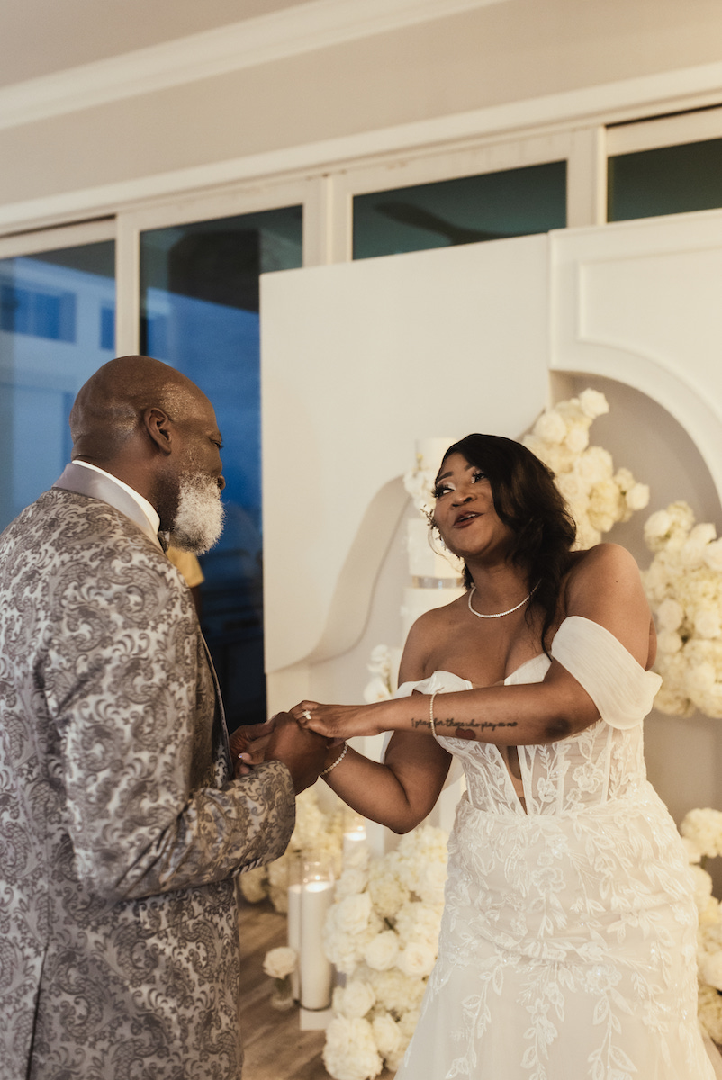 A bride in a white dress holds hands and smiles at a man in a patterned suit jacket, standing in front of a floral wedding backdrop.