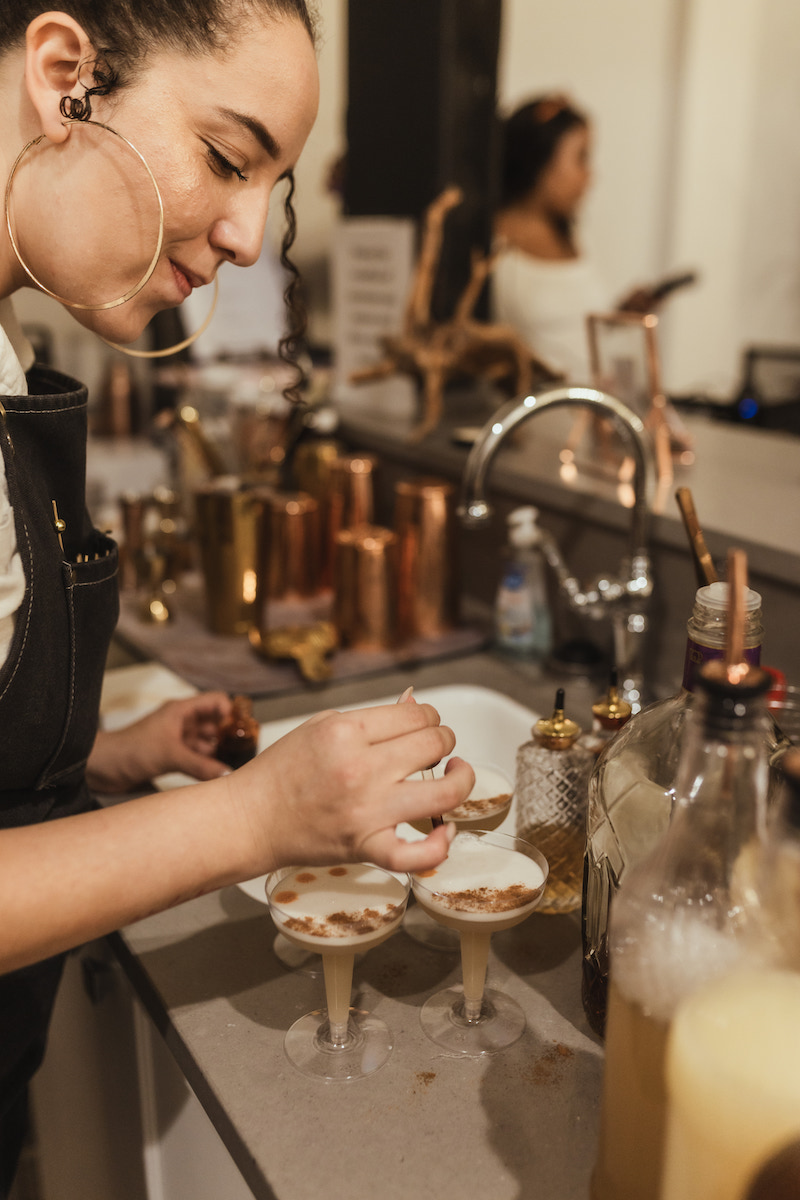 A woman behind a bar adds garnish to cocktails in coupe glasses, with bottles and bar tools visible on the counter.
