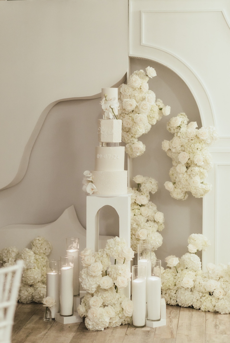 A white wedding cake on a pedestal surrounded by white flowers and candles, with a decorative white backdrop.