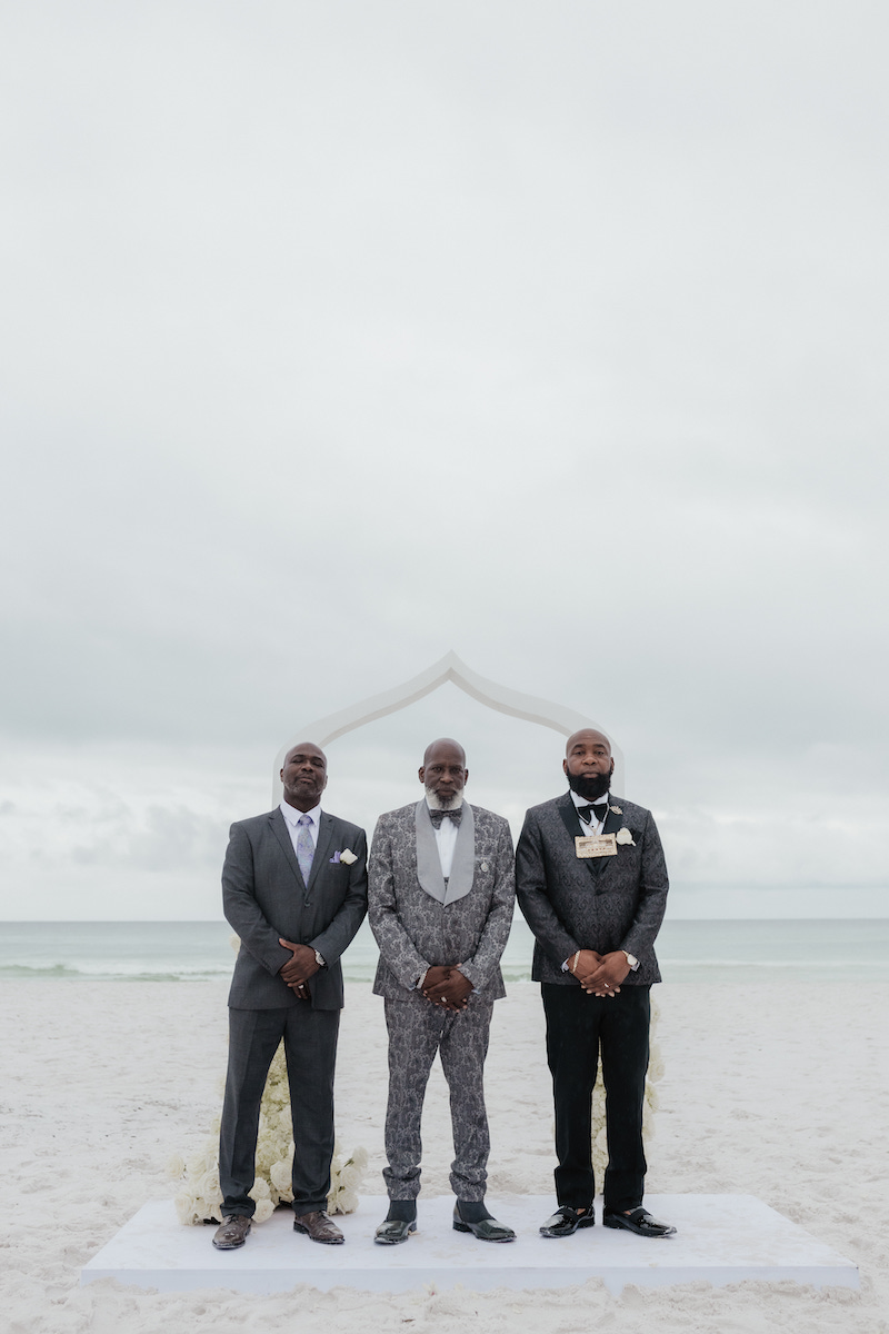 Three men in suits stand side by side on a beach under an overcast sky, with a simple arch in the background.
