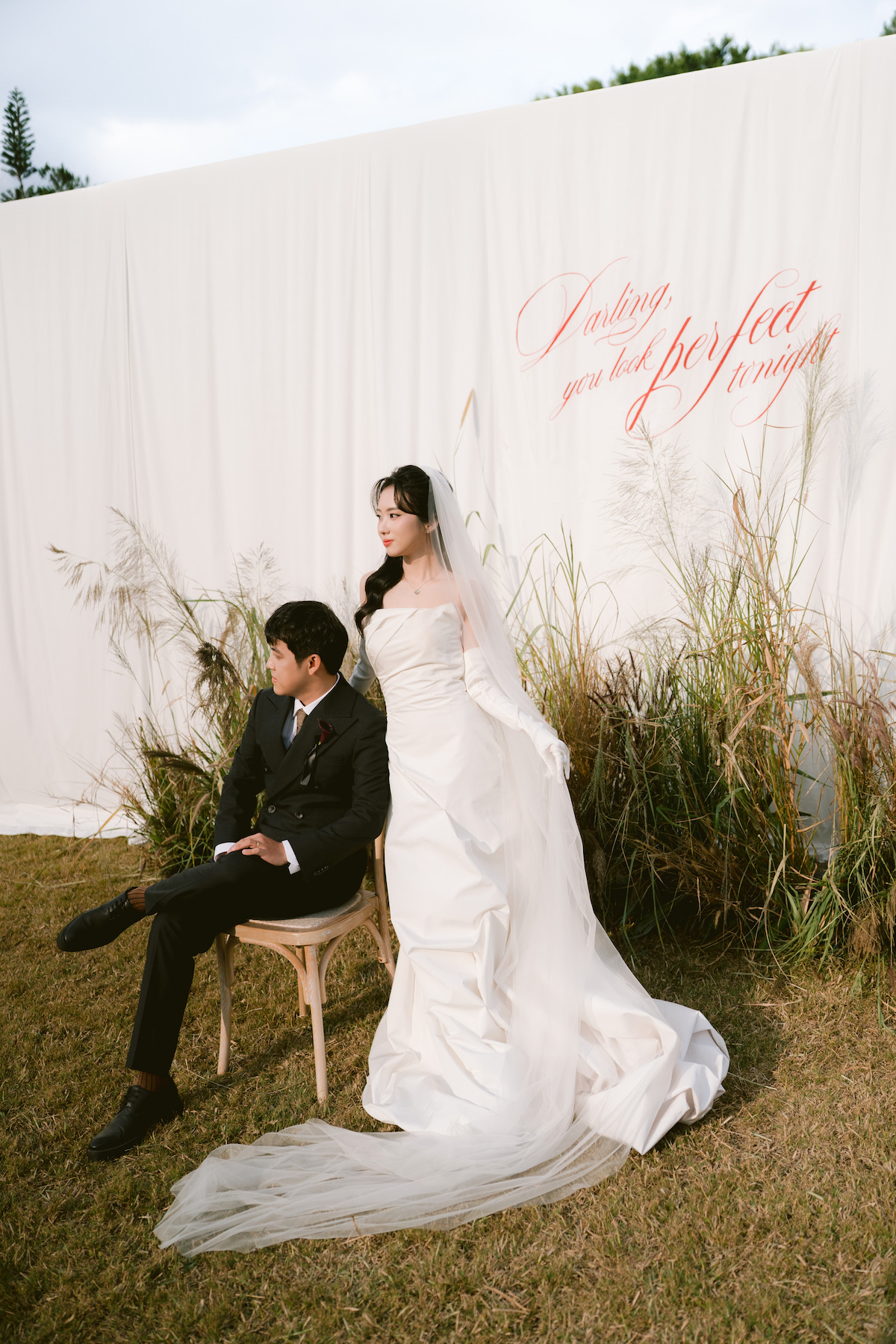 A bride in a white gown stands beside a groom seated on a chair outdoors, with a white backdrop and dried grass decorations behind them.