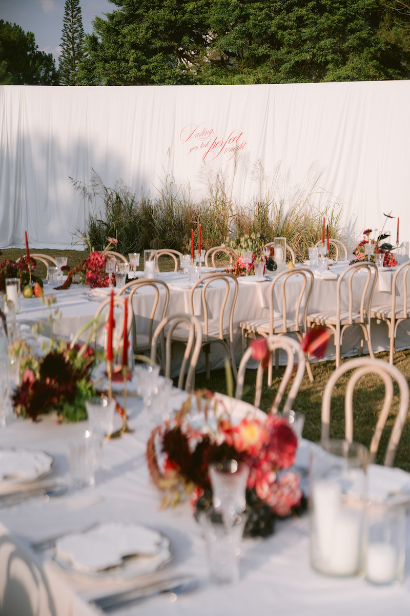 Outdoor dining area with white tablecloths, beige chairs, red candles, floral centerpieces, and a white backdrop with greenery and text, set on a grassy lawn.