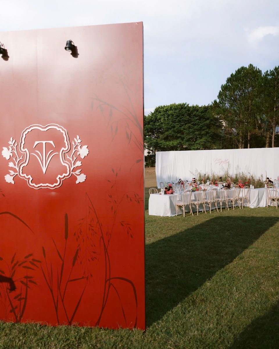 A large red panel with a white decorative emblem stands on grass near a long outdoor dining table set with white linens and chairs. Trees and a white backdrop are in the background.