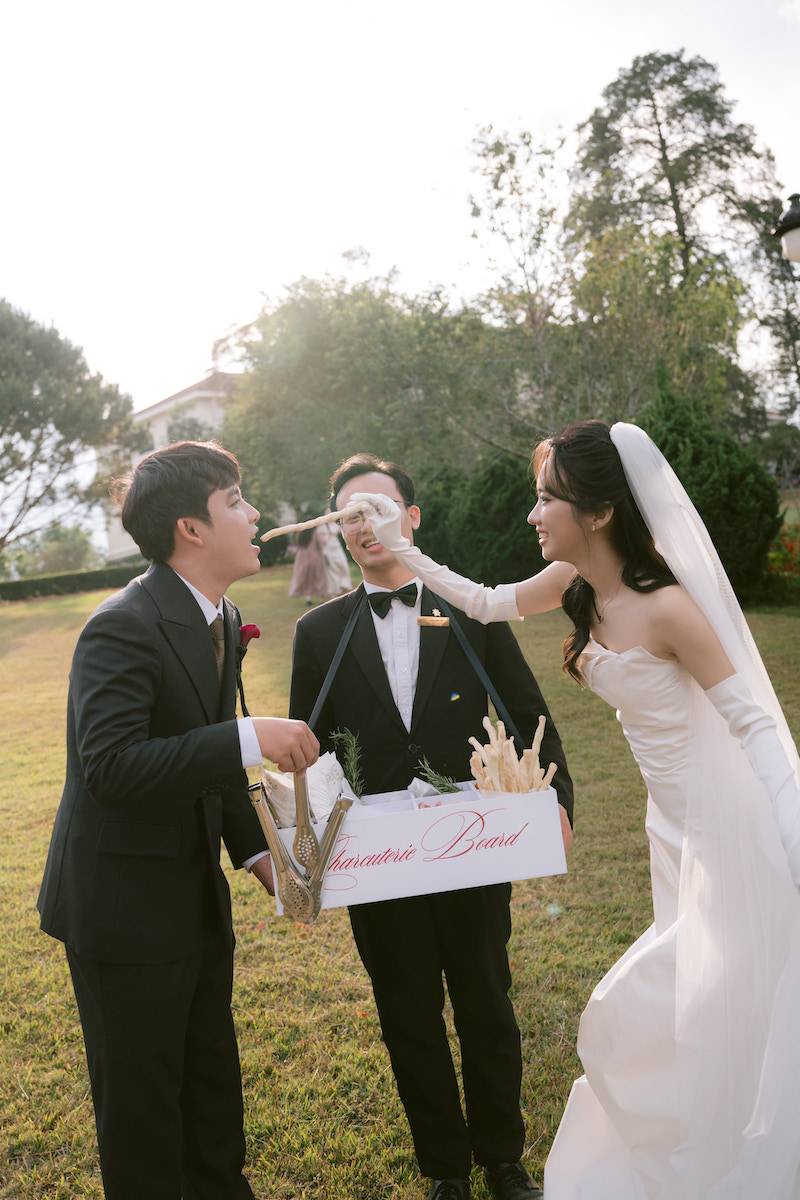 A bride in a white dress feeds a groom breadsticks, while another man in a suit holds a box labeled "homemade bread" outdoors.