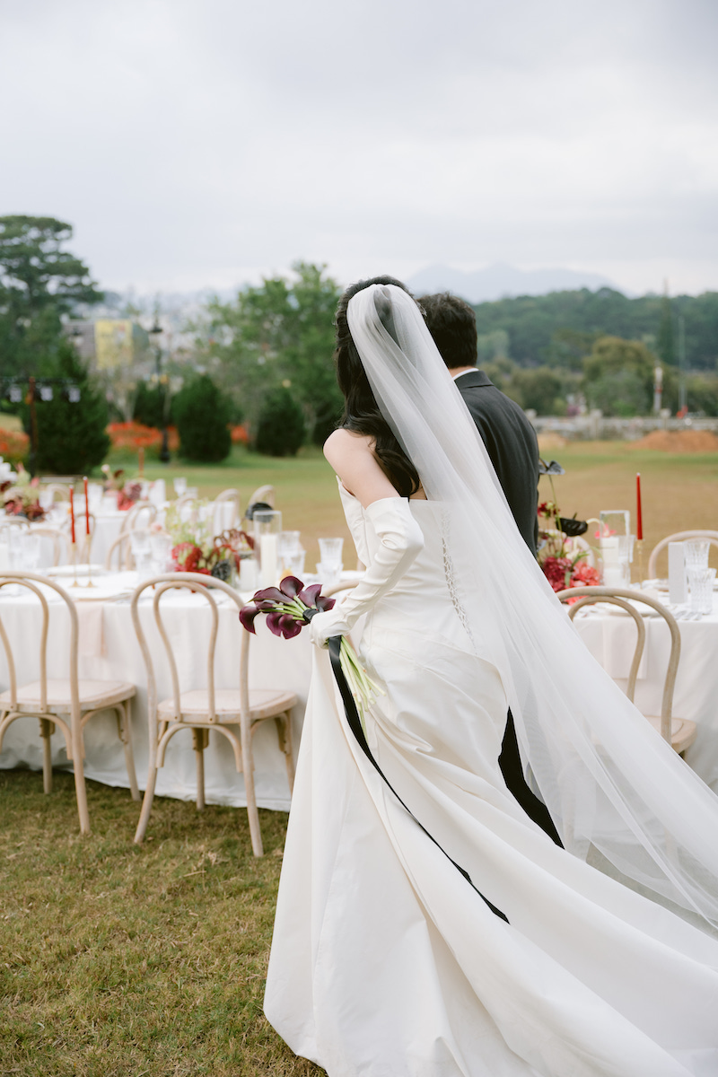A bride and groom walk together outdoors beside a long, decorated table set for a wedding reception, with greenery and cloudy sky in the background.