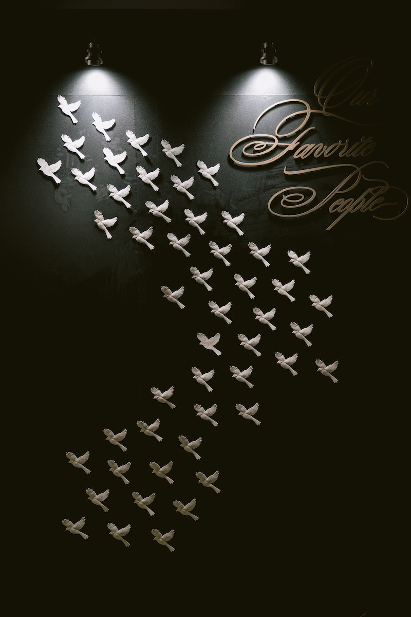 Dozens of white bird silhouettes arranged on a dark wall with two spotlights above and cursive writing on the upper right side.